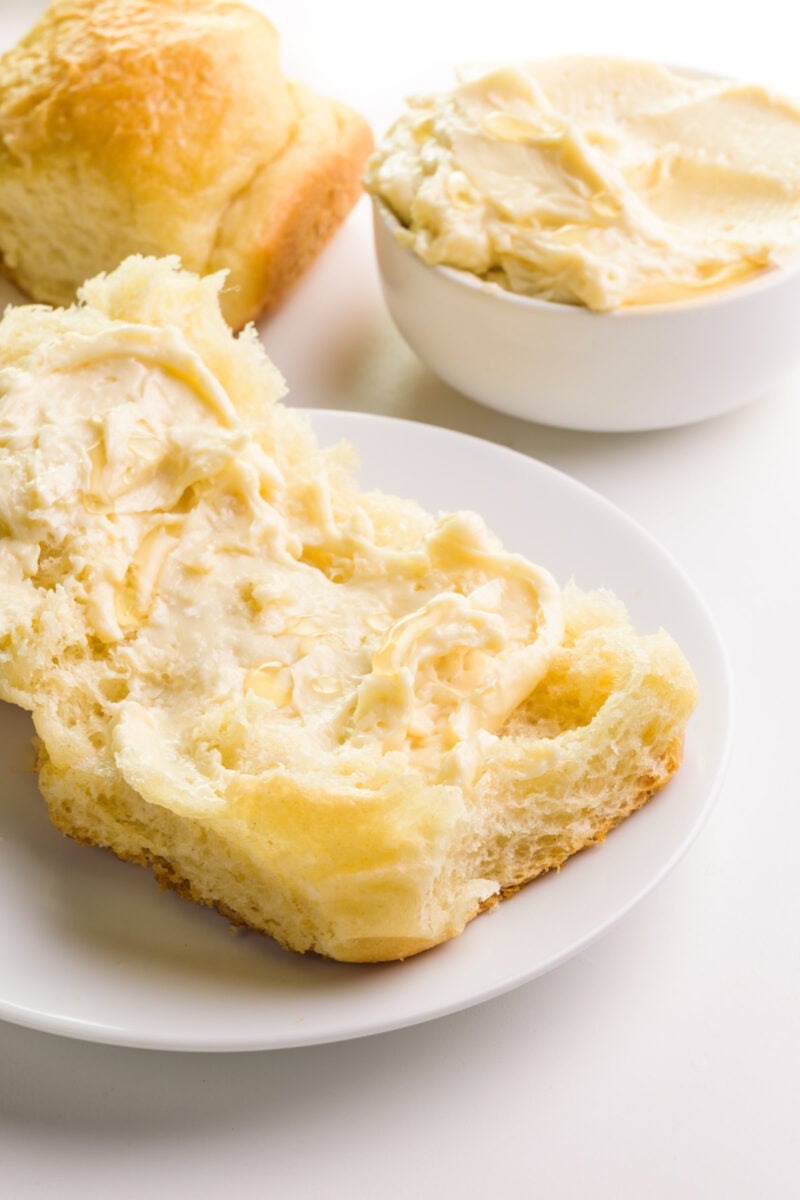 Vegan honey butter is spread over a dinner rolls. A bowl with more honey butter and dinner rolls are in the background.