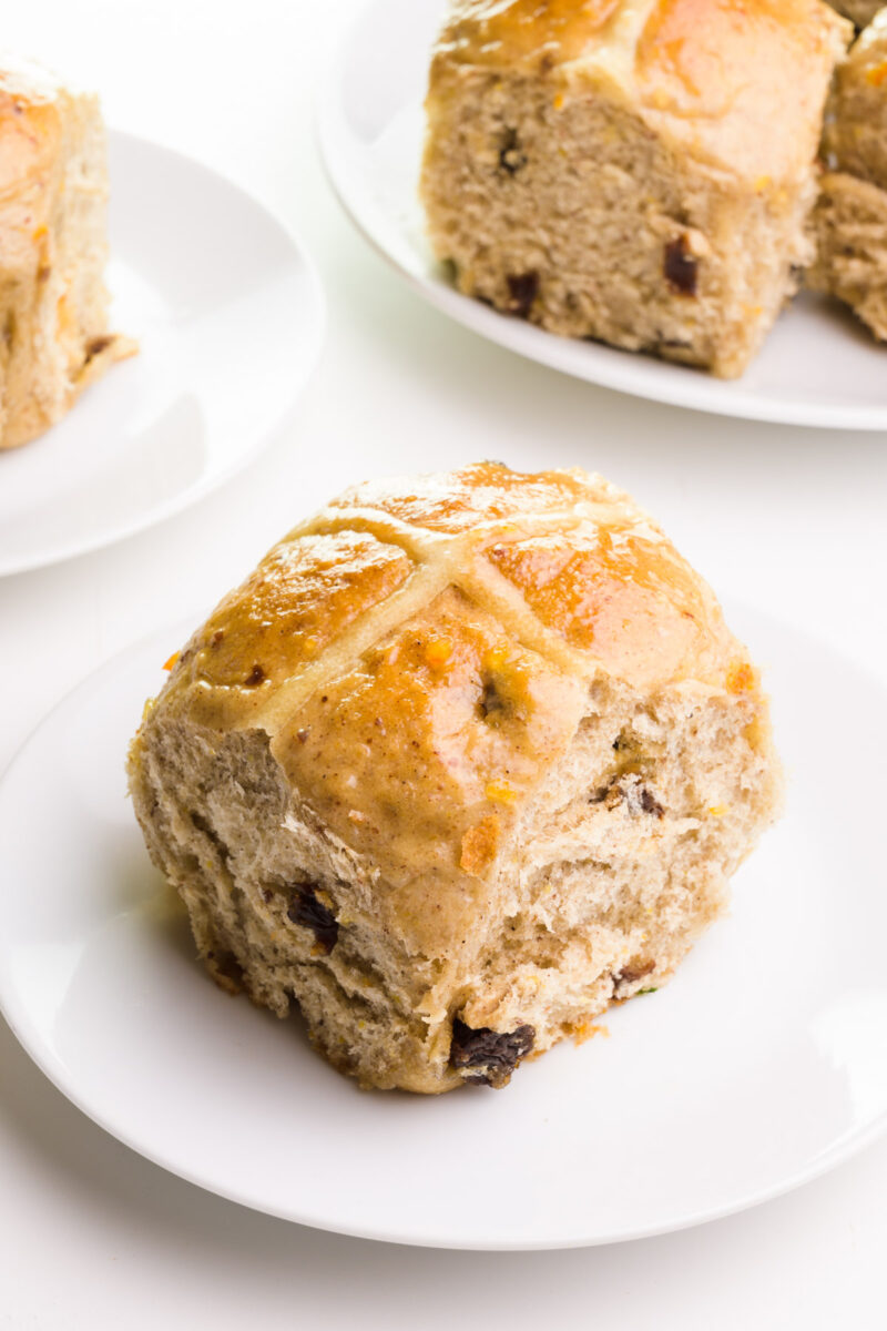 A vegan hot cross bun sits on a plate. There are more buns on plates in the background.