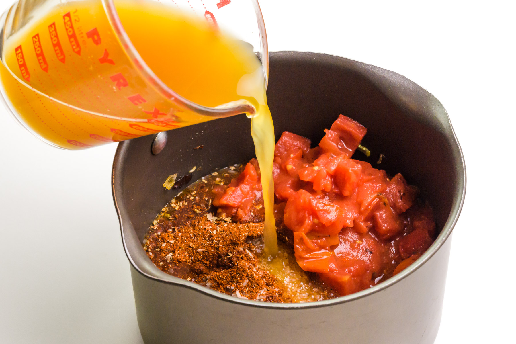 Vegetable broth is being poured into a saucepan with diced tomatoes, spices, and more.