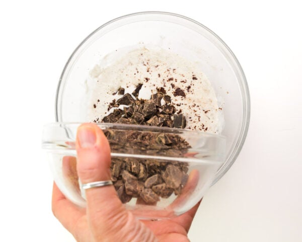 A hand holds a bowl of chopped chocolate, pouring it into a bowl with heated vegan creamer.