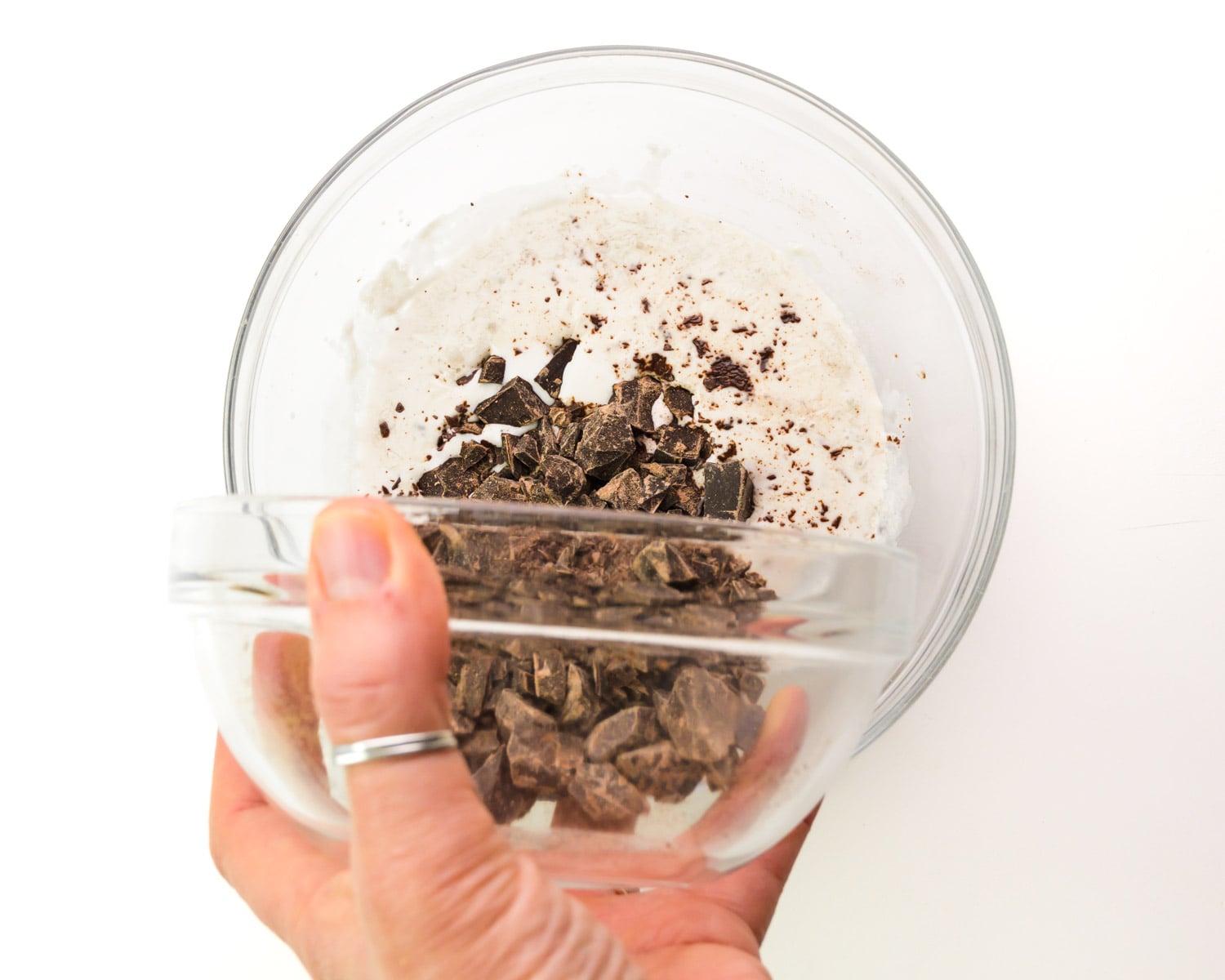 A hand holds a bowl of chopped chocolate, pouring it into a bowl with heated vegan creamer.