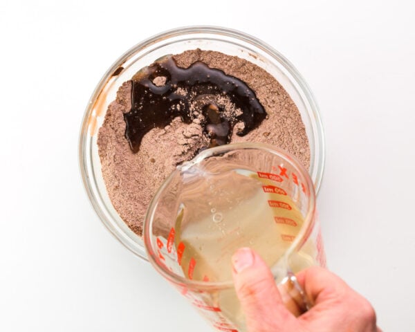 A hand holds a glass measuring cup full of liquid ingredients and is pouring it into a bowl with a flour and cocoa powder mixture.