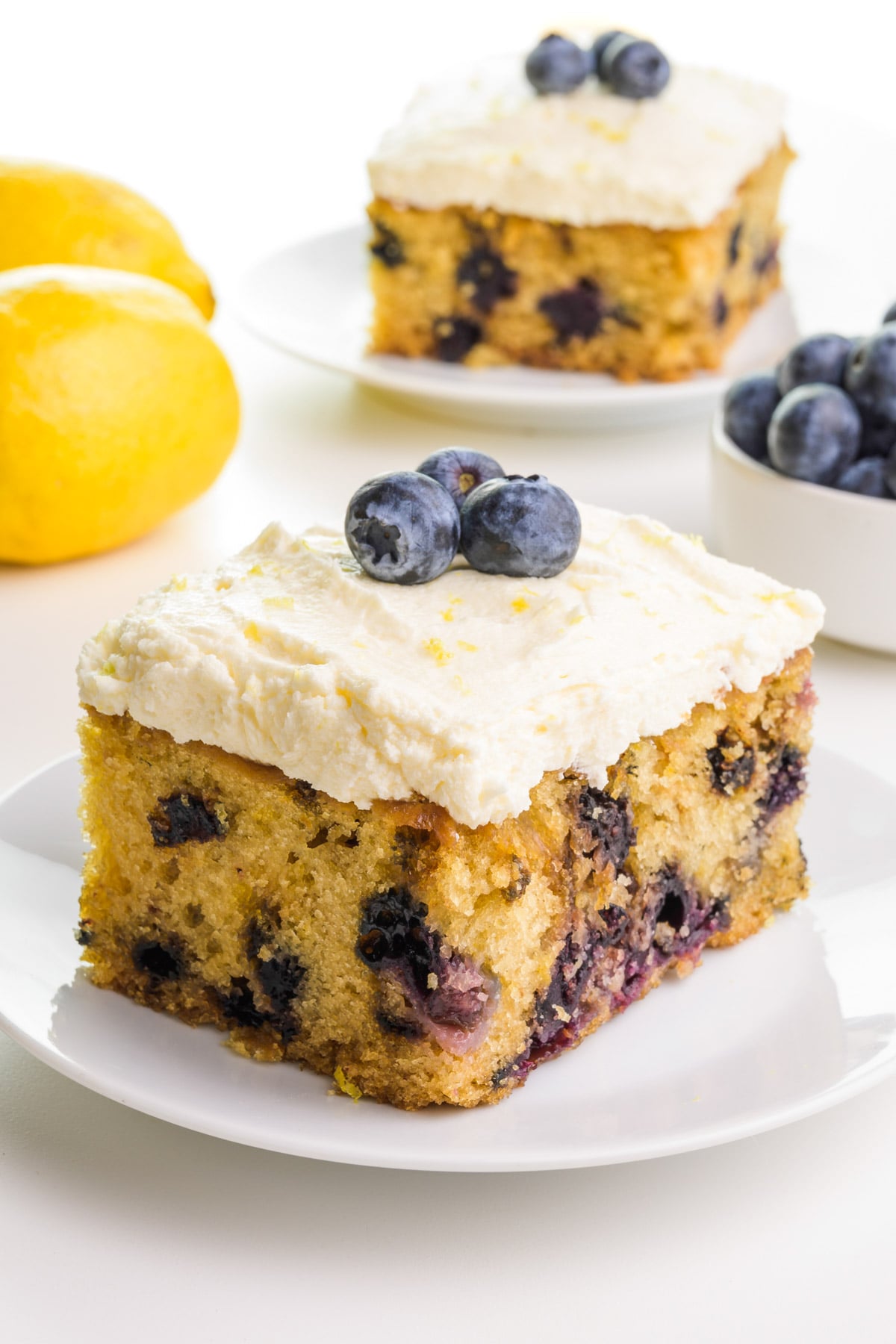 A lemon blueberry cake has frosting on top and fresh blueberries. There is a lemon in the background, another slice of cake, and a bowl of fresh blueberries.
