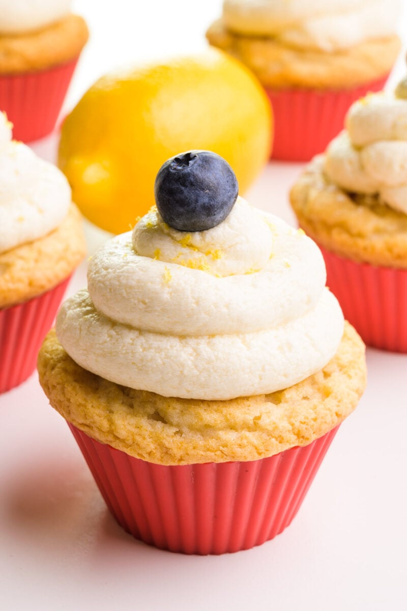 A cupcake has vegan lemon frosting on top with a blueberry and lemon zest. There are more cupcakes and a lemon in the background.