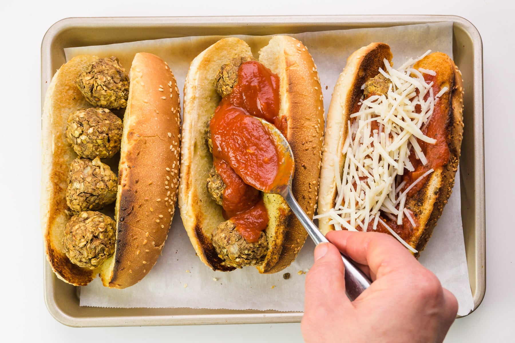 A hand holds a spoon, drizzling marinara sauce over vegan sandwiches.