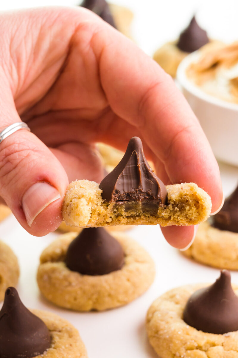 A hand holds a vegan Hershey kiss cookie with a bite taken out next to more cookies in the background.
