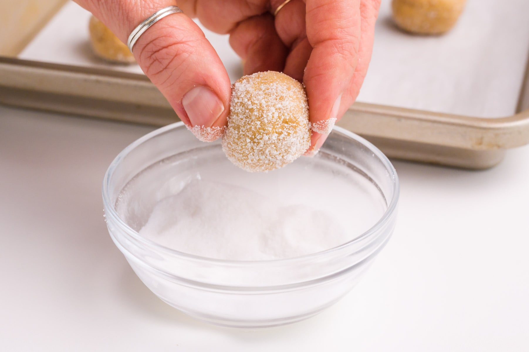 A hand holds a cookie dough bowl over a bowl of sugar. A baking pan is in the background.