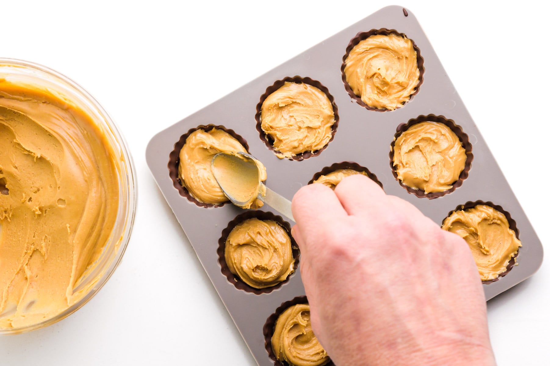 A hand holds a spoon pressing peanut butter over set chocolate in candy molds.