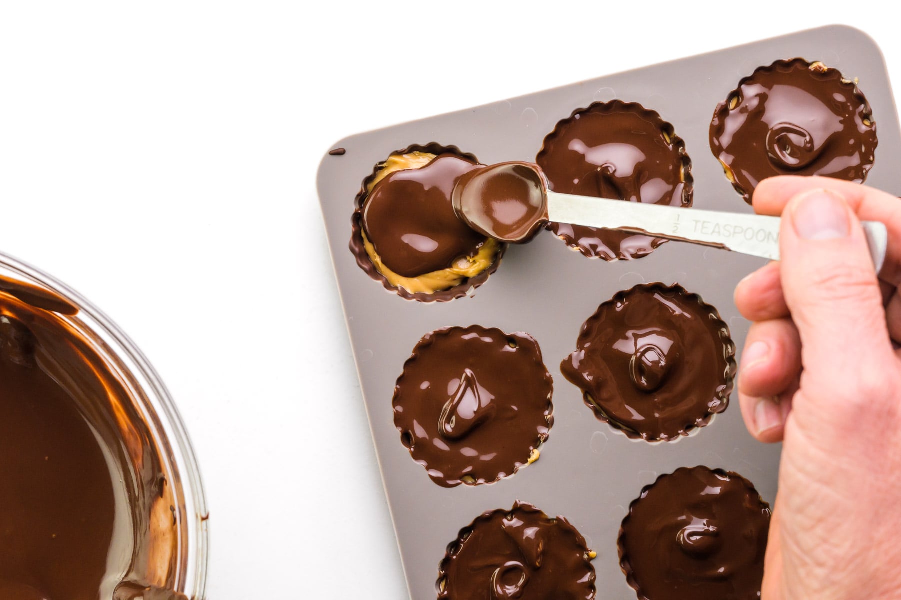 A hand holds a spoon, spreading meted chocolate over candy molds.