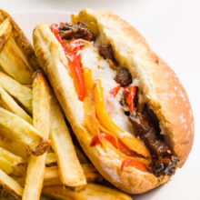 A closeup of a vegan Philly cheesesteak sandwich sitting next to French fries on a plate.