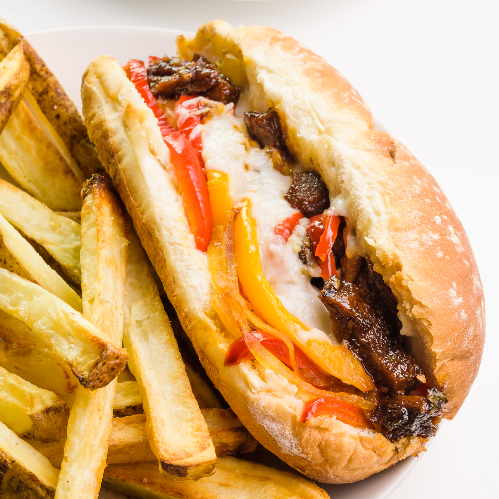 A closeup of a vegan Philly cheesesteak sandwich sitting next to French fries on a plate.