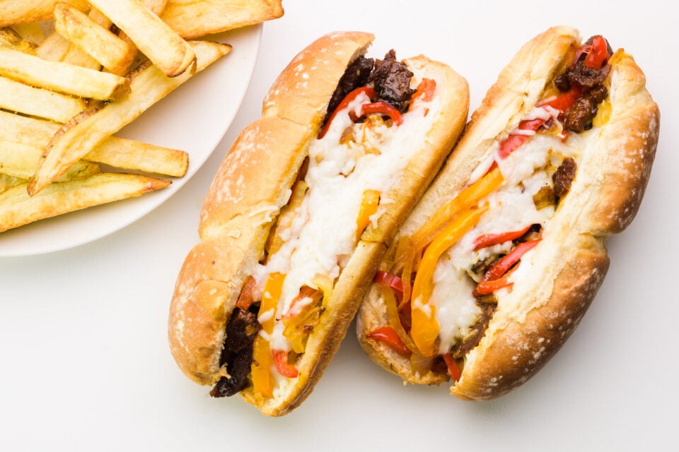 Looking down on two vegan cheesesteak sandwiches sitting next to a plate of fries.