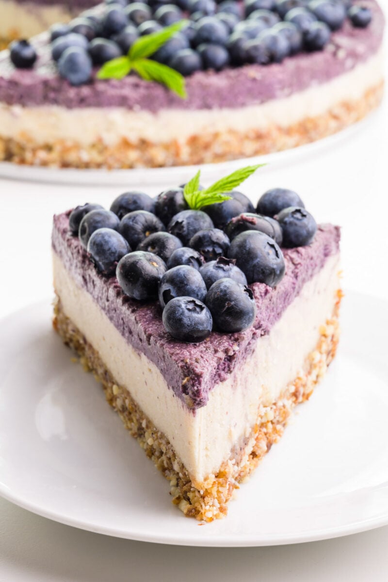 A slice of raw cheesecake has blueberries on top. It's sitting on a white plate. The rest of the cheesecake is in the background.