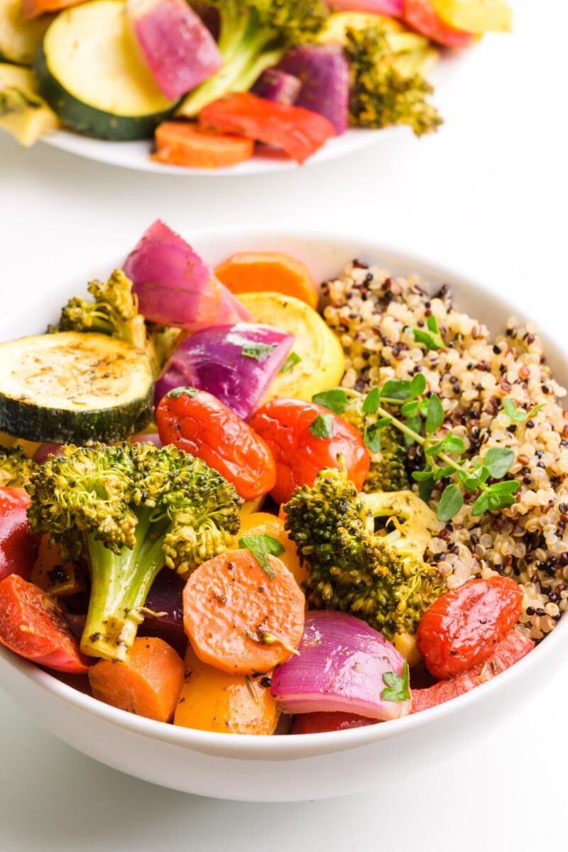 A bowl of vegan roasted veggies has quinoa on the side. There is a plate of vegetables in the background.