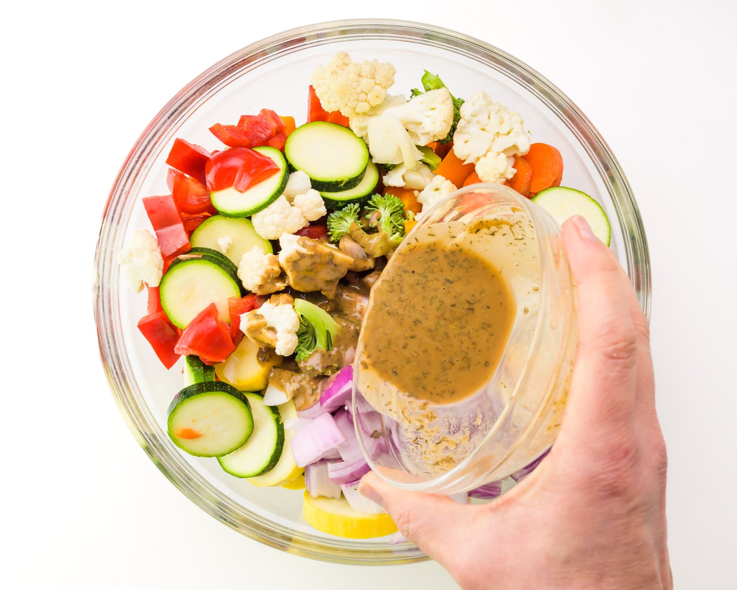 A hand holds a bowl of sauce, pouring it over chopped veggies in a bowl.