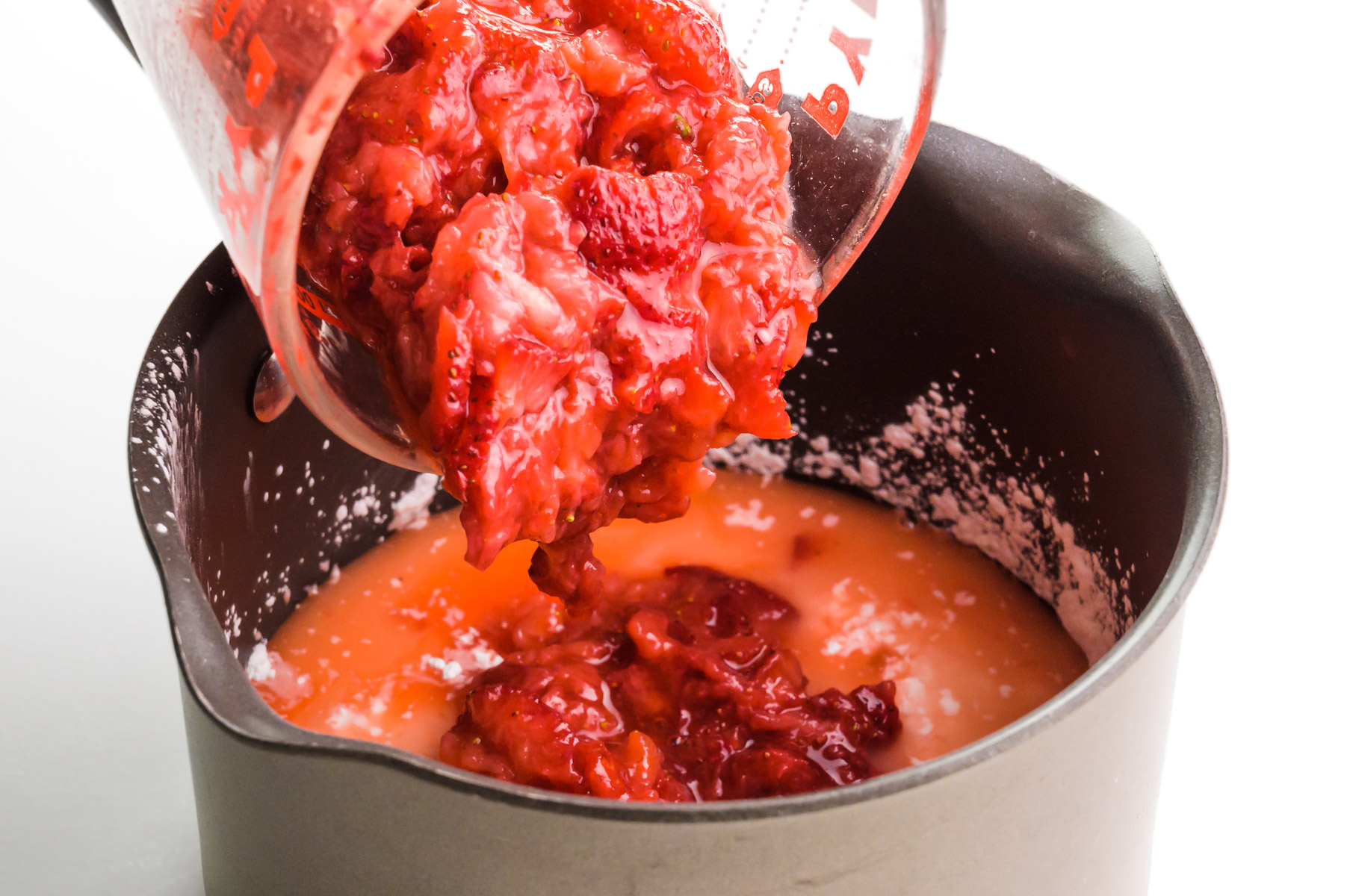 Mashed strawberries are being poured from a bowl into a saucepan.