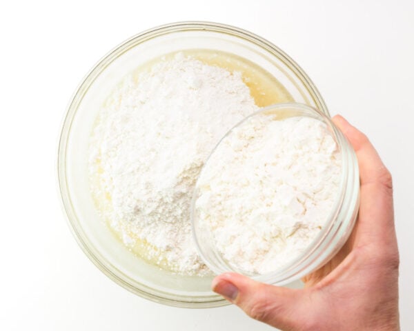 A hand pours a bowl of flour into a larger bowl with other ingredients.