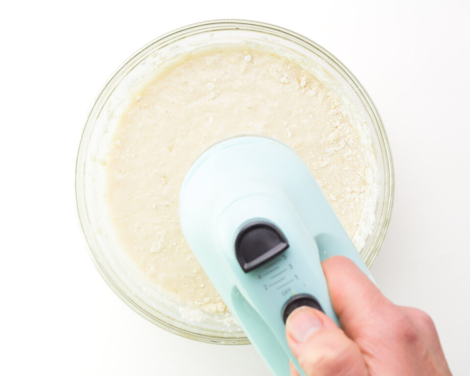 A hand holds a mixer, using it to mix cake batter in a mixing bowl.