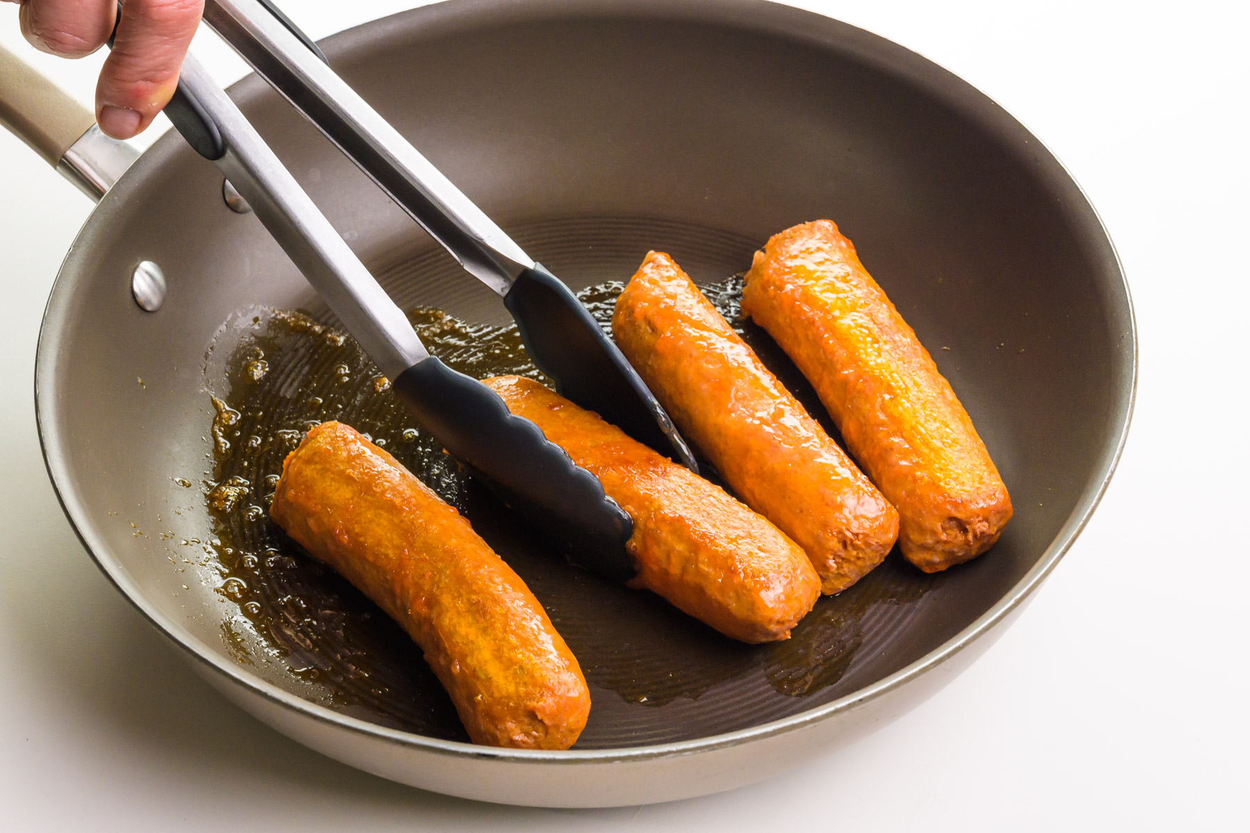 A hand holds tongs, turning vegan sausages in a skillet.