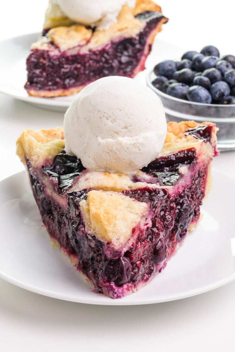 A slice of vegan berry pie sits on a plate. It has ice cream on top. There is another slice behind it and a bowl of fresh blueberries.