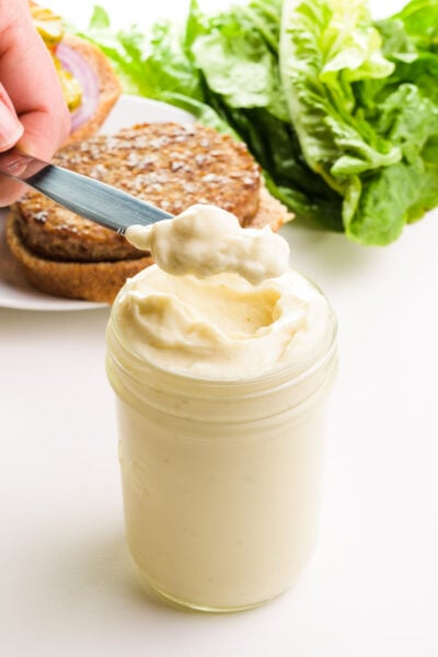 A hand holds a scoop of mayo on a knife, hovering over a jar full of it. There is a veggie burger and lettuce in the background.