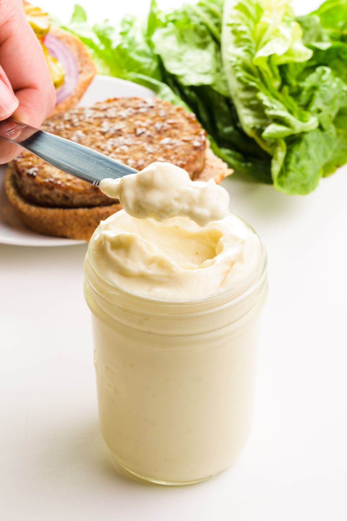 A hand holds a scoop of mayo on a knife, hovering over a jar full of it. There is a veggie burger and lettuce in the background.