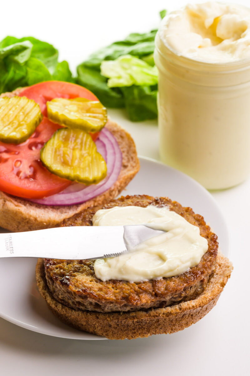 Vegan mayo is being slathered over a veggie burger. The rest of the bun has chopped onions, tomatoes, and pickles on top. The jar of mayo and lettuce are in the background.