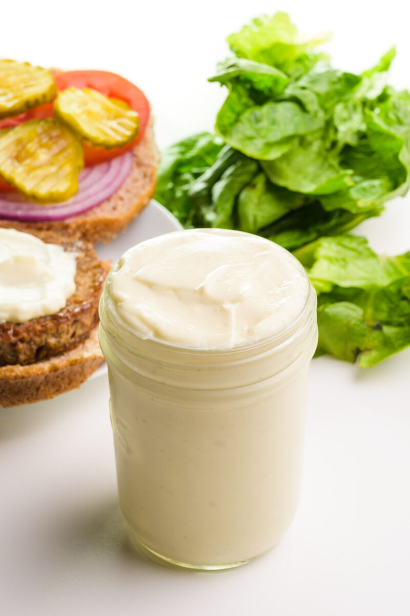 Jar of mayonnaise with lettuce and a sandwich topped with pickles, tomatoes, and onion in the background.