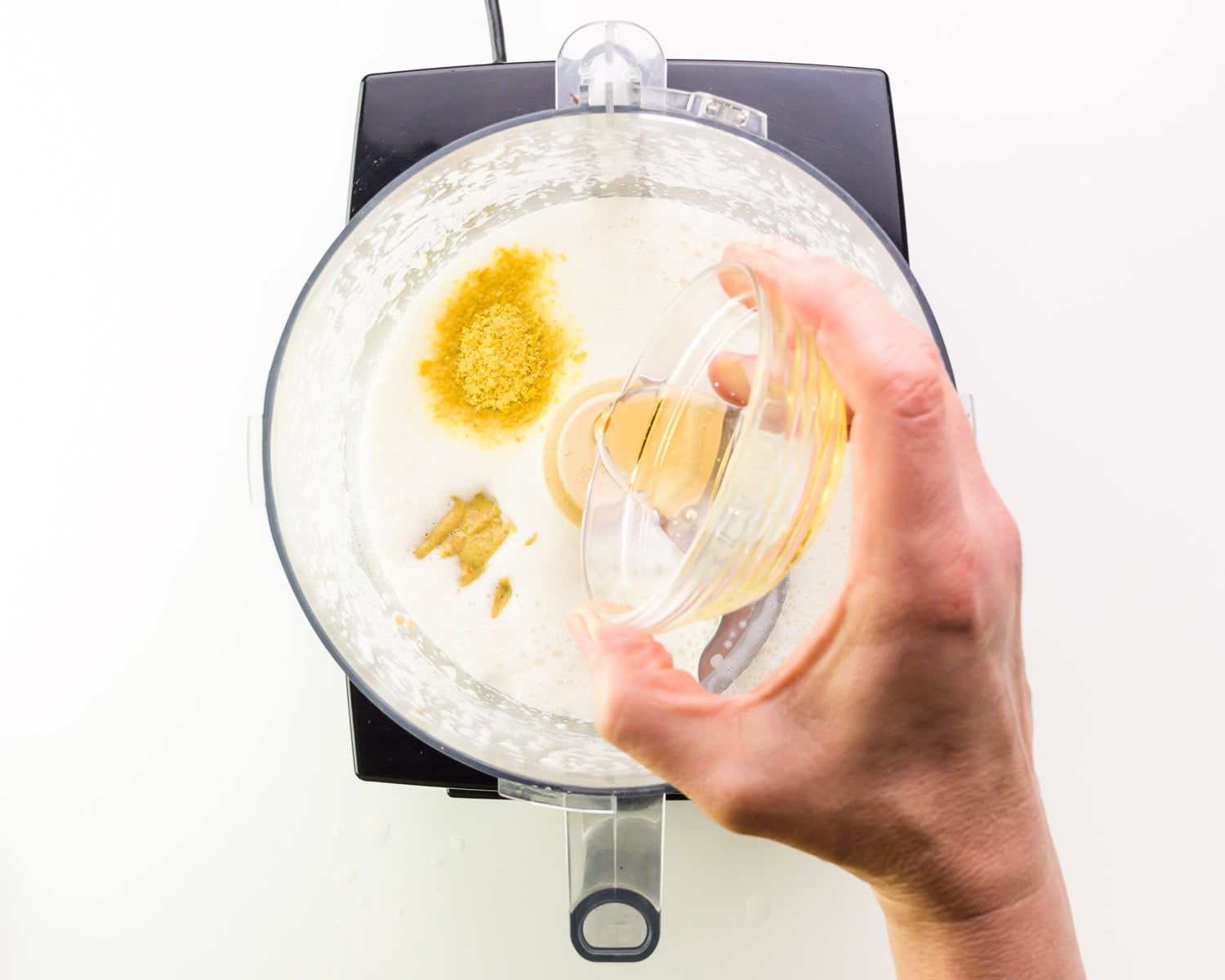 A hand holds a bowl of agave nectar, pouring it into a food processor with other ingredients.