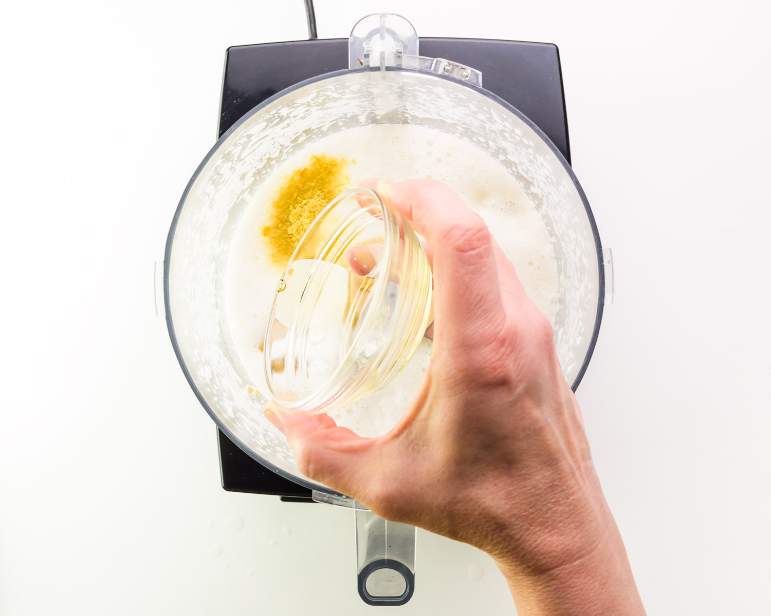 A hand holds a jar of vinegar, pouring it into a food processor with other ingredients.