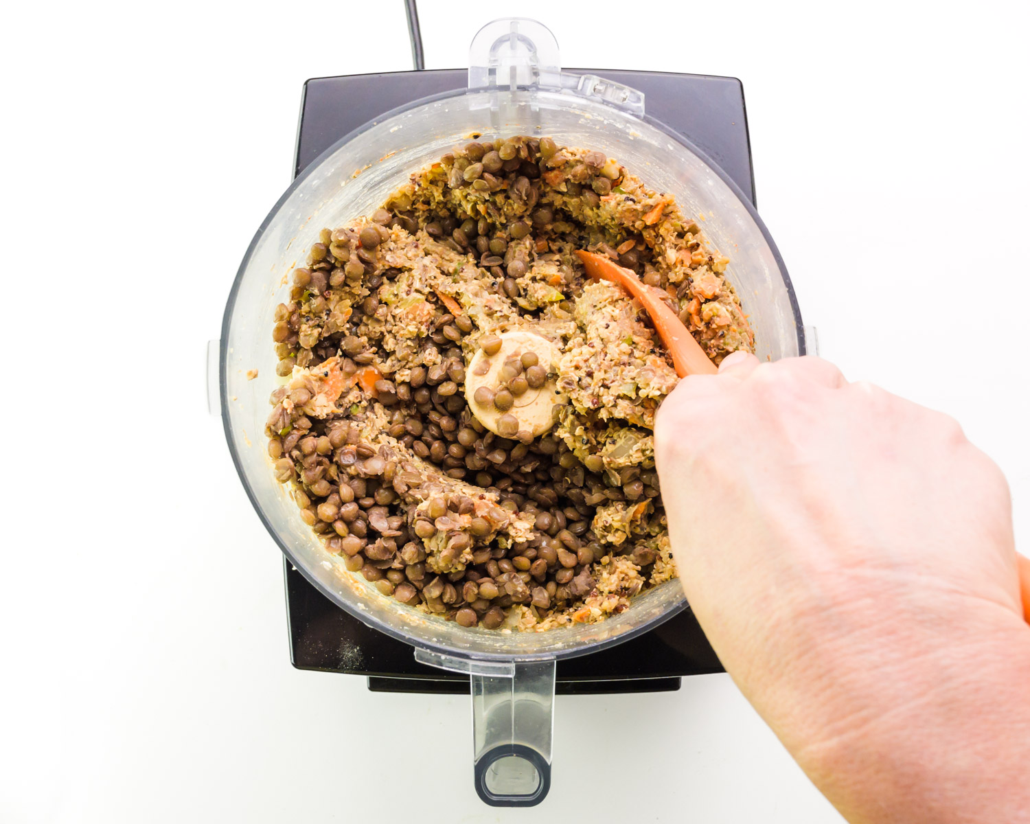 A hand holds a rubber spatula, stirring ingredients in a food processor.