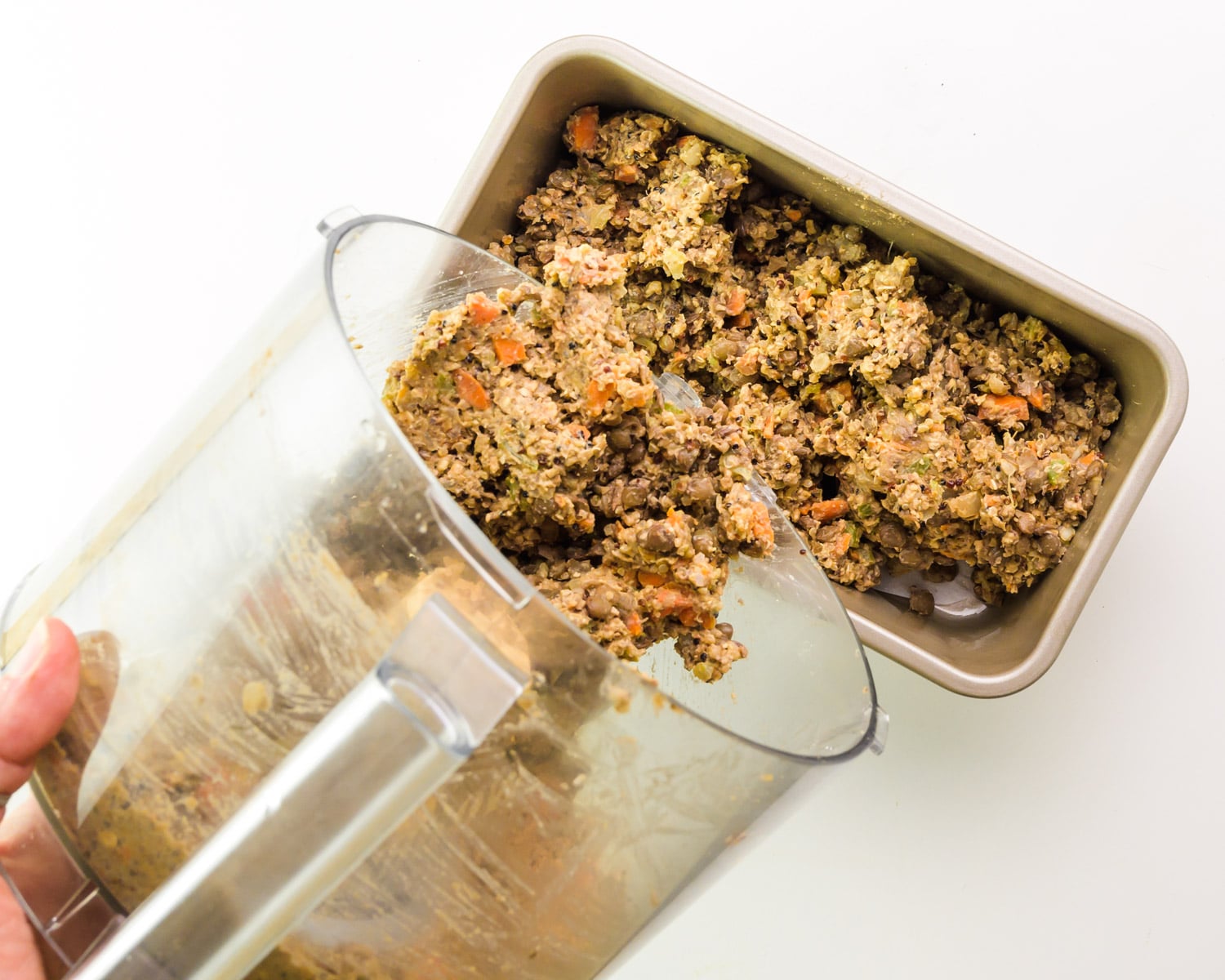 A bean mixture is being poured from a food processor bowl into a loaf pan.