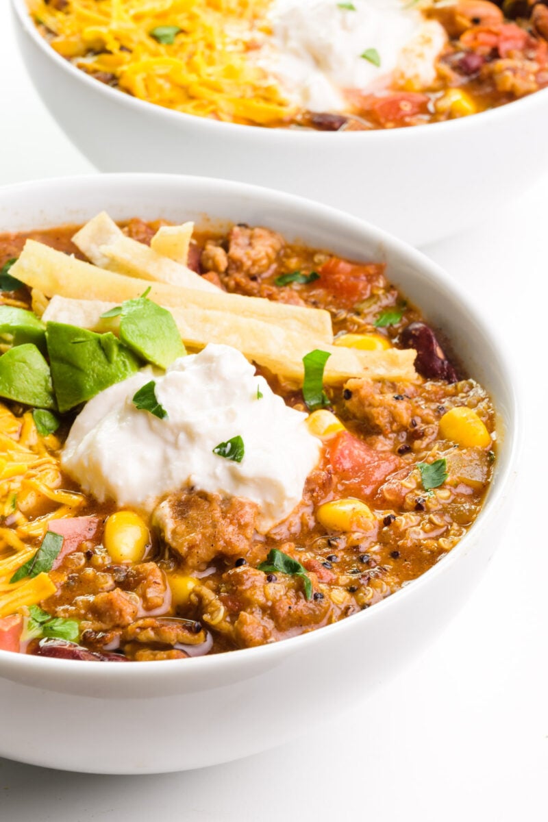 A bowl of vegan taco soup has vegan sour cream, melted vegan cheese, avocados, and tortilla strips on top. There's another bowl in the background.
