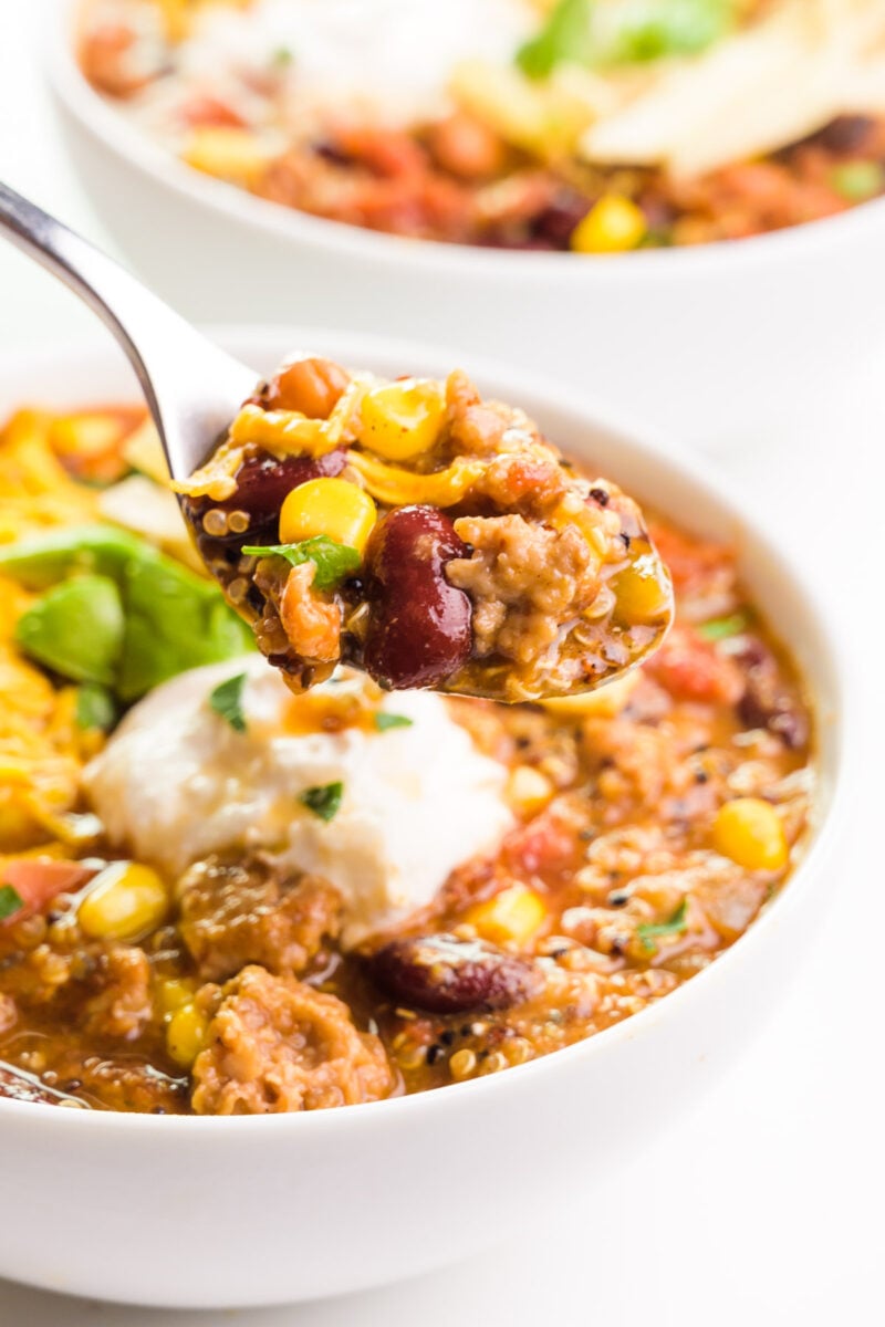 A spoon holds soup and is hovering over a bowl with more soup.
