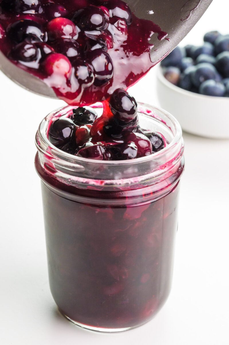Blueberry glaze is being poured from a saucepan into a glass jar. There are fresh blueberries in the background.