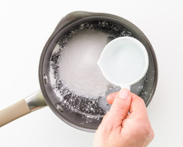 A hand holds a measuring cup of water, adding it to a saucepan with sugar and cornstarch.