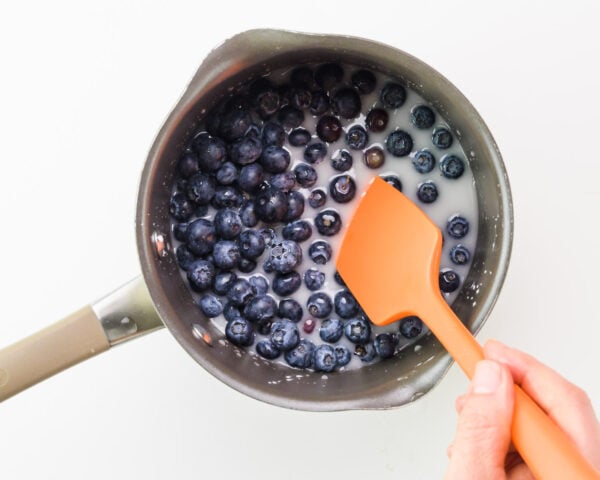 A hand holds an orange spatula, using it to stir blueberries in a saucepan.