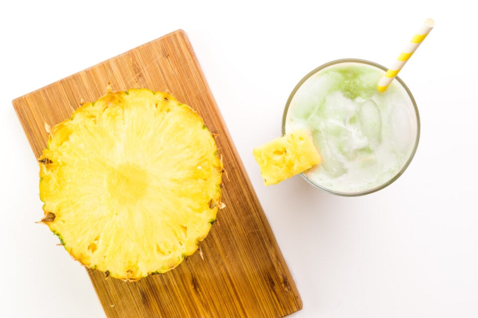 Looking down on a cut pineapple on a cutting board. It sits beside a glass with iced matcha drink inside it.