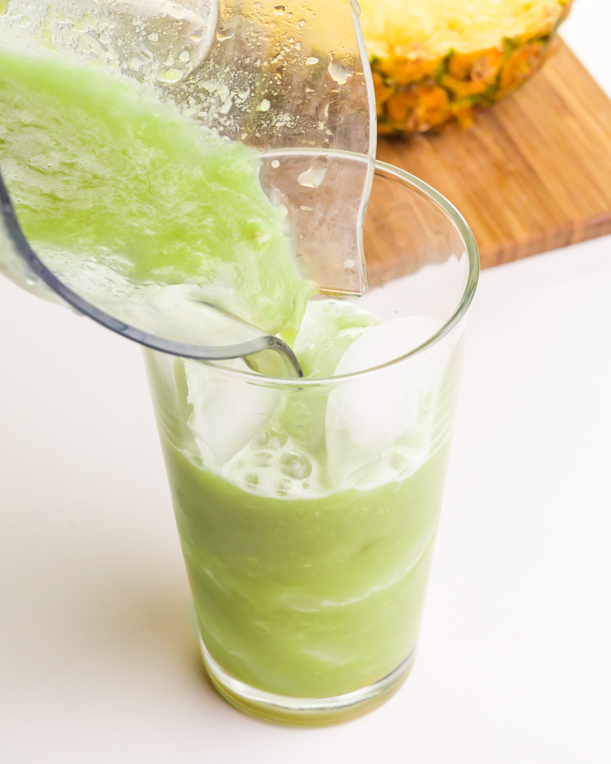 Matcha syrup is being poured into a glass with ice. There is sliced pineapple on a cutting board in the background.