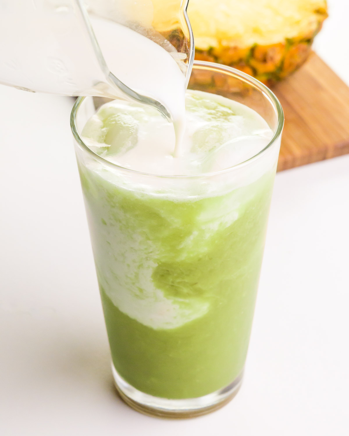 Coconut milk is being poured into a glass with a matcha mixture. There is fresh pineapple on a cutting board in the background.