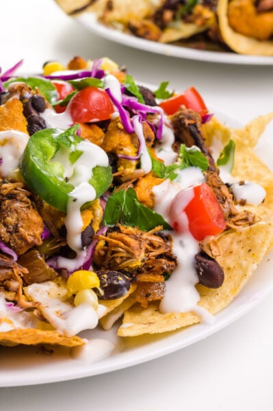 A plate of jackfruit nachos has chopped jalapeños, sour cream, and more as toppings. There's another plate in the background.