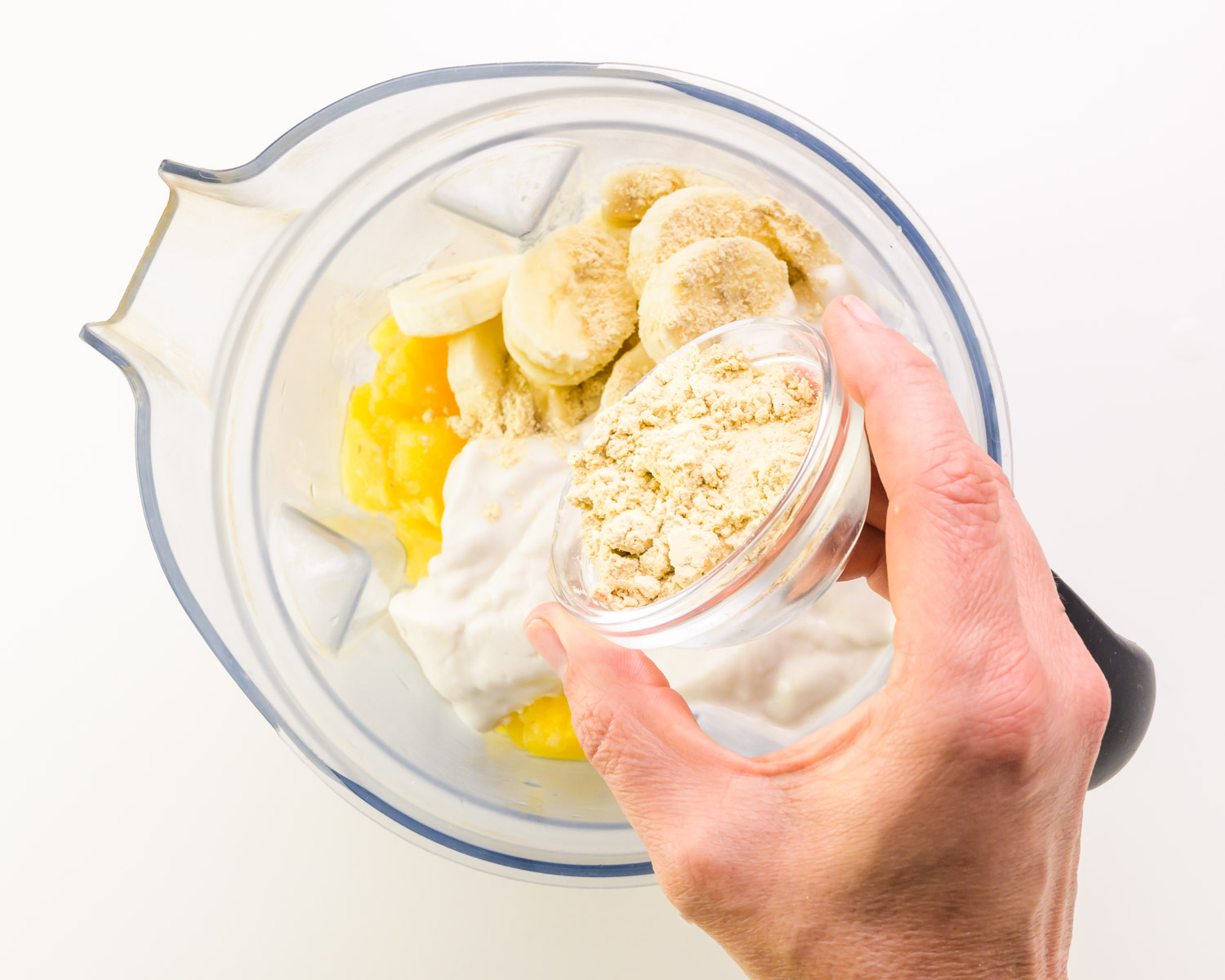 Vegan yogurt is being added to a blender bowl with other ingredients.
