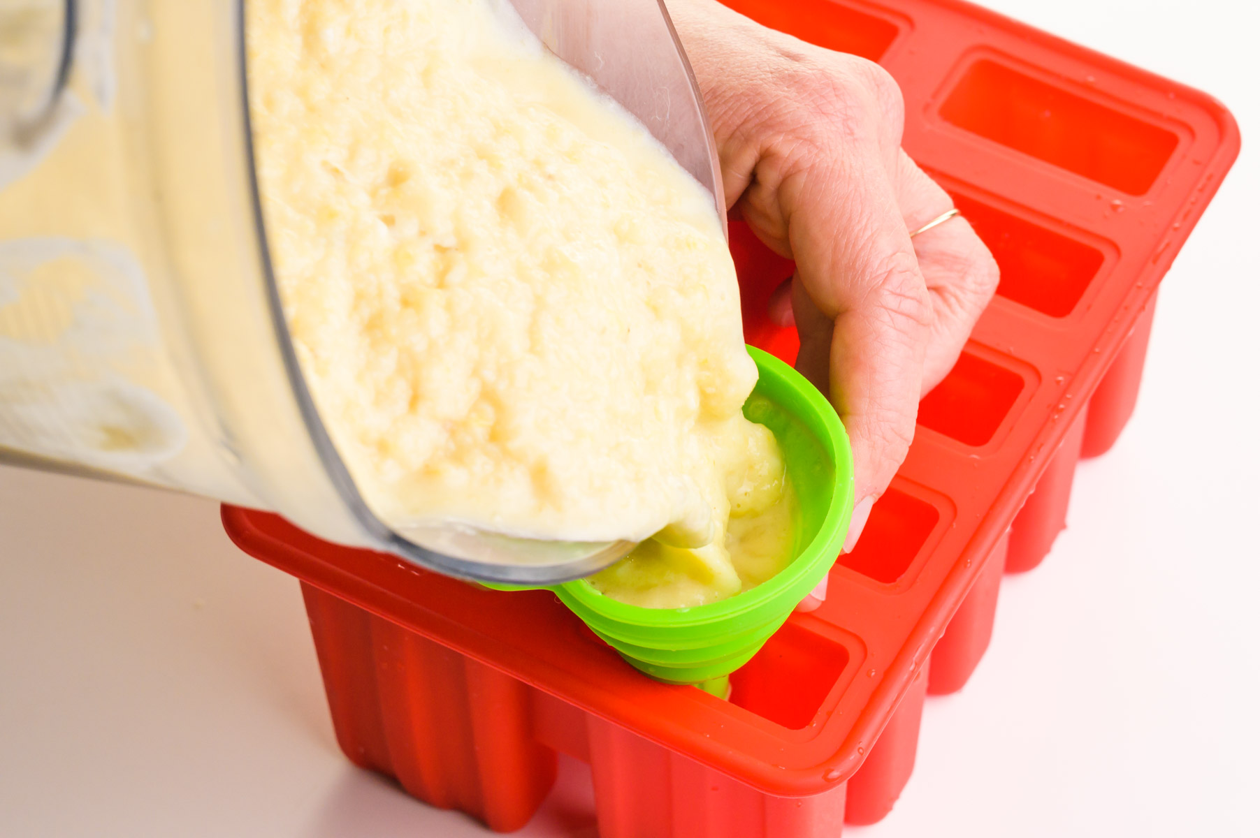 A creamy mixture is being poured from a blender into a funnel, filling up popsicles in a mold.