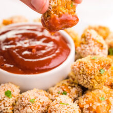 A hand holds a sweet potato tater tot dipped in ketchup, dangling over a plate with more tater tots and a bowl of ketchup.