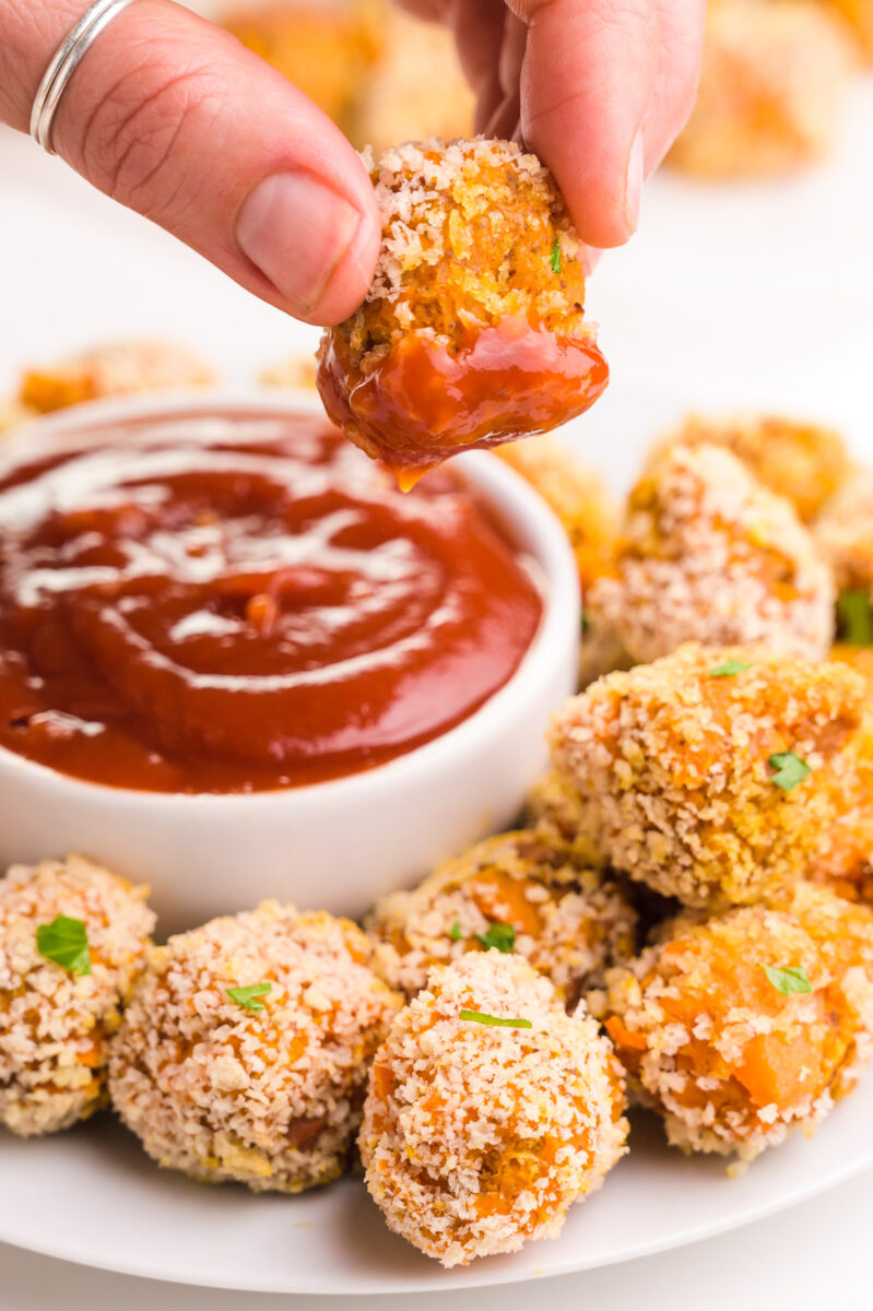 A hand holds a sweet potato tater tot dipped in ketchup, dangling over a plate with more tater tots and a bowl of ketchup.