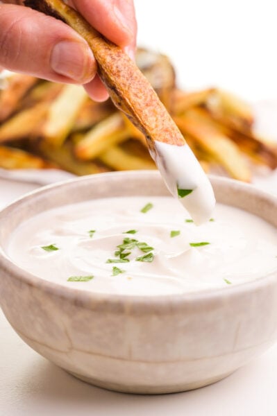 A hand holds a French fry, recently dipped in vegan aioli, hovering over a bowl of it. There are more fries in the background.