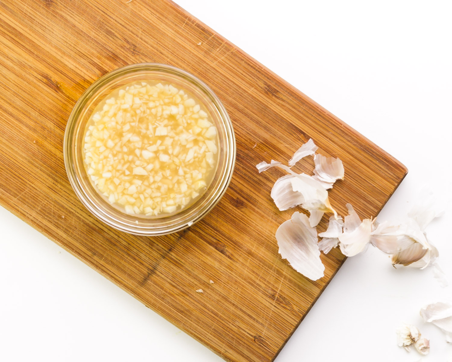 Chopped garlic sits in lemon juice in a bowl on a cutting board. There is chopped garlic beside it.