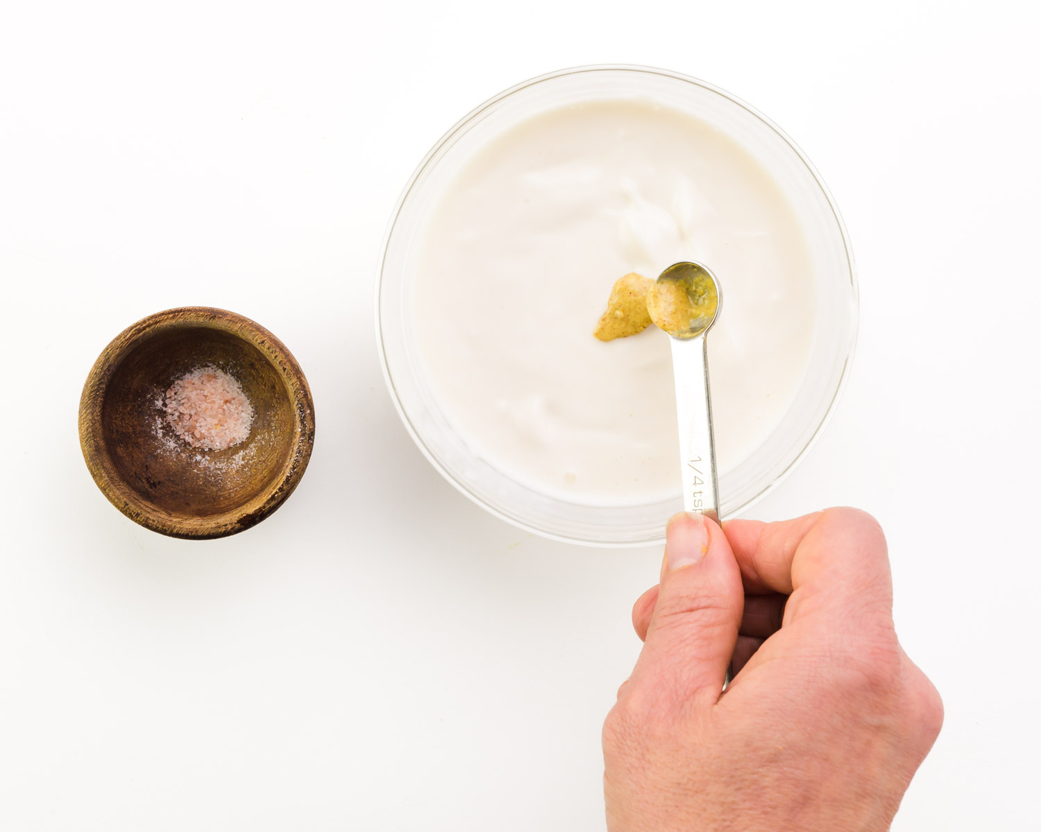 Brown mustard is being added to a bowl of aioli. There is a wooden bowl with seal salt beside it.