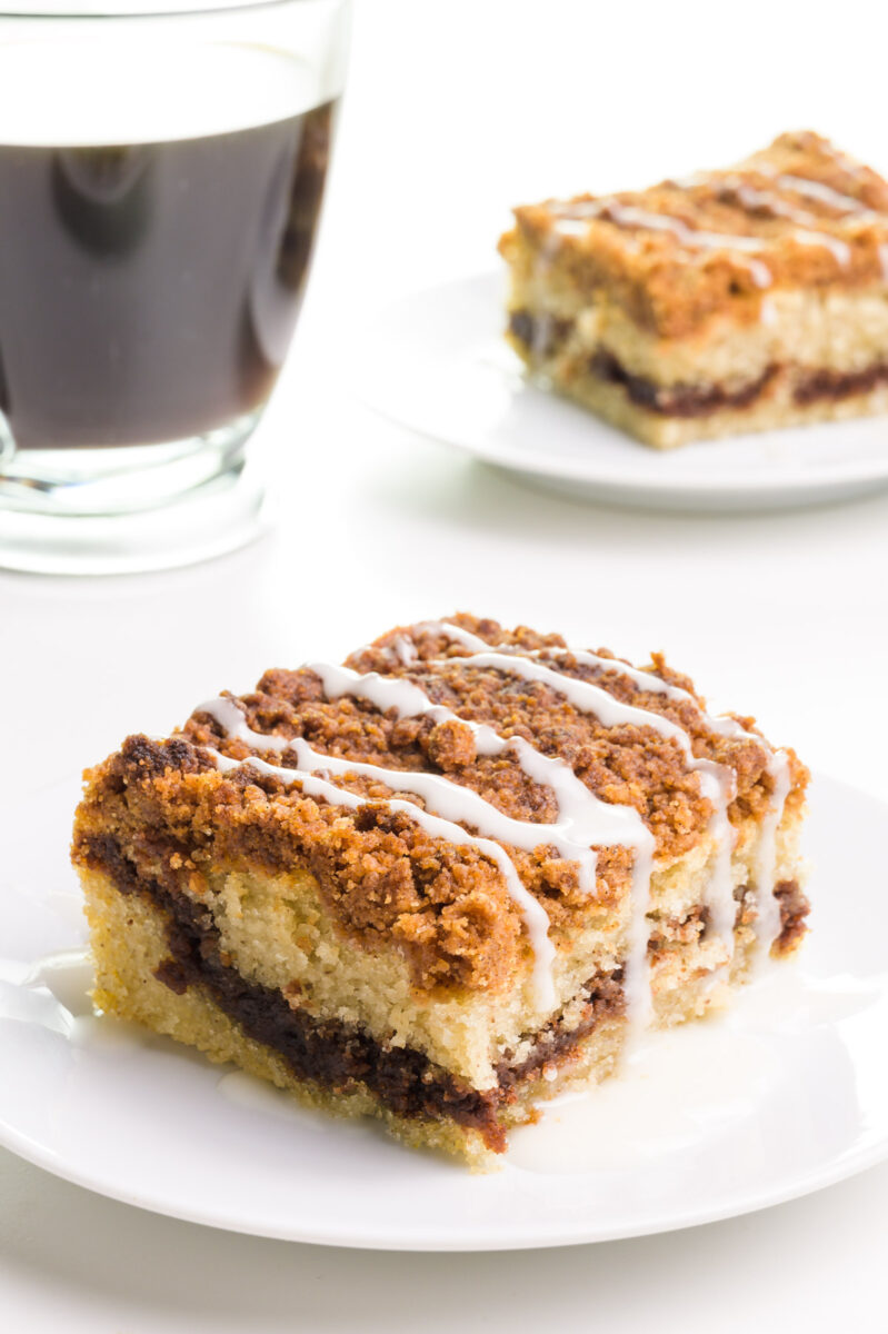 A slice of coffee cake on a plate sits in front of another slice and a cup of coffee.