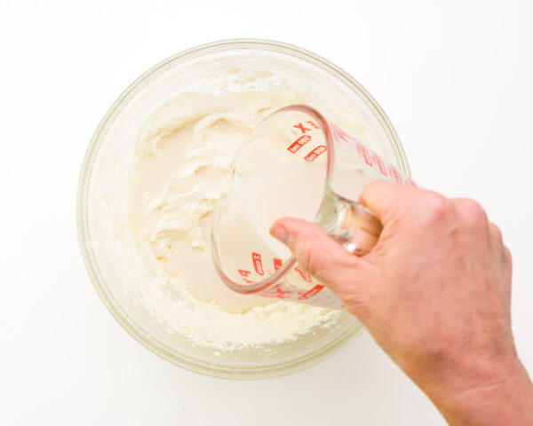 A hand holds a measuring glass full of plant-based milk pouring it into a bowl with whipped vegan butter.